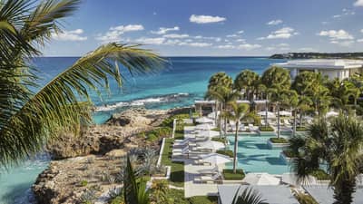 Looking down over Anguilla outdoor pool, patio, large rocks along blue-green ocean waterfront, blue sky